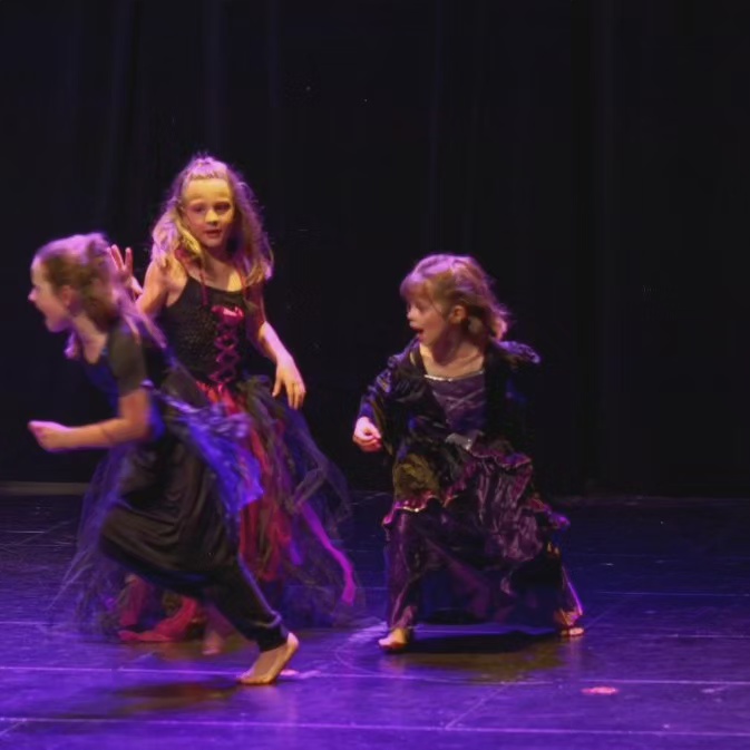 Trois jeunes danseuses en costumes sombres, en mouvement sur scène sous une lumière violette.
