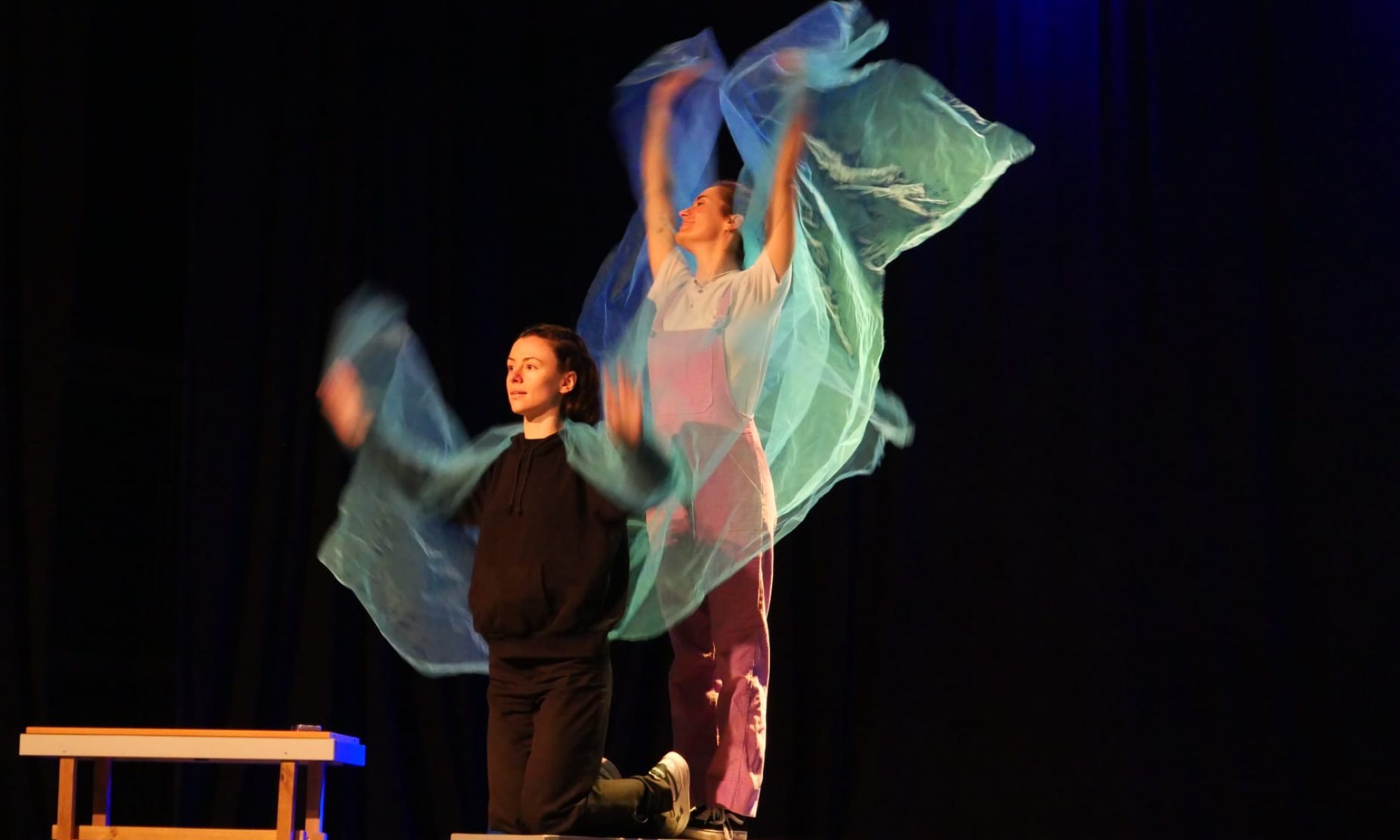 Deux comédiens sur scène, l'un debout sur un banc les bras levés dans un voile bleu translucide, dans le spectacle Toi t'es moche