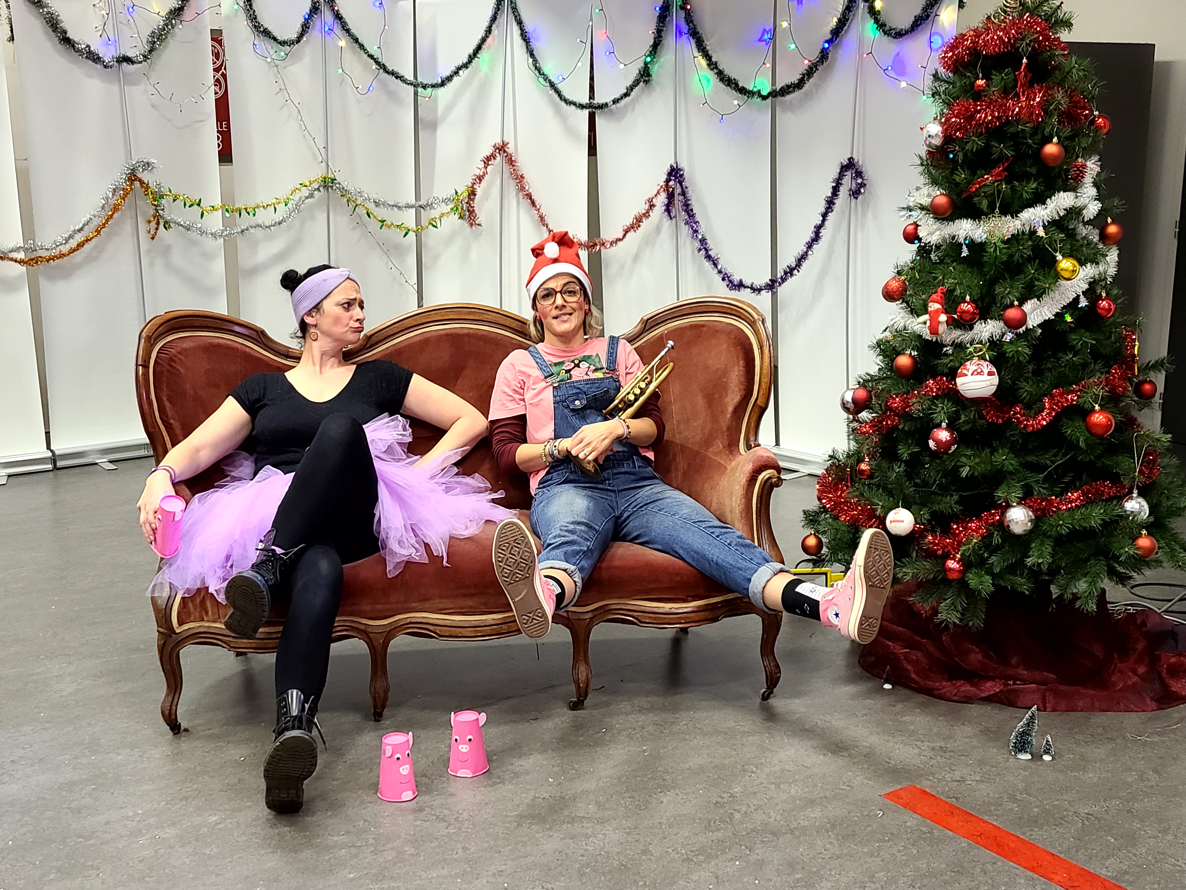 Deux comédiennes assises sur un canapé rouge devant le sapin de Noël : l'une en tutu lilas, l'autre en bonnet de Père Noël, les jambes allongées.