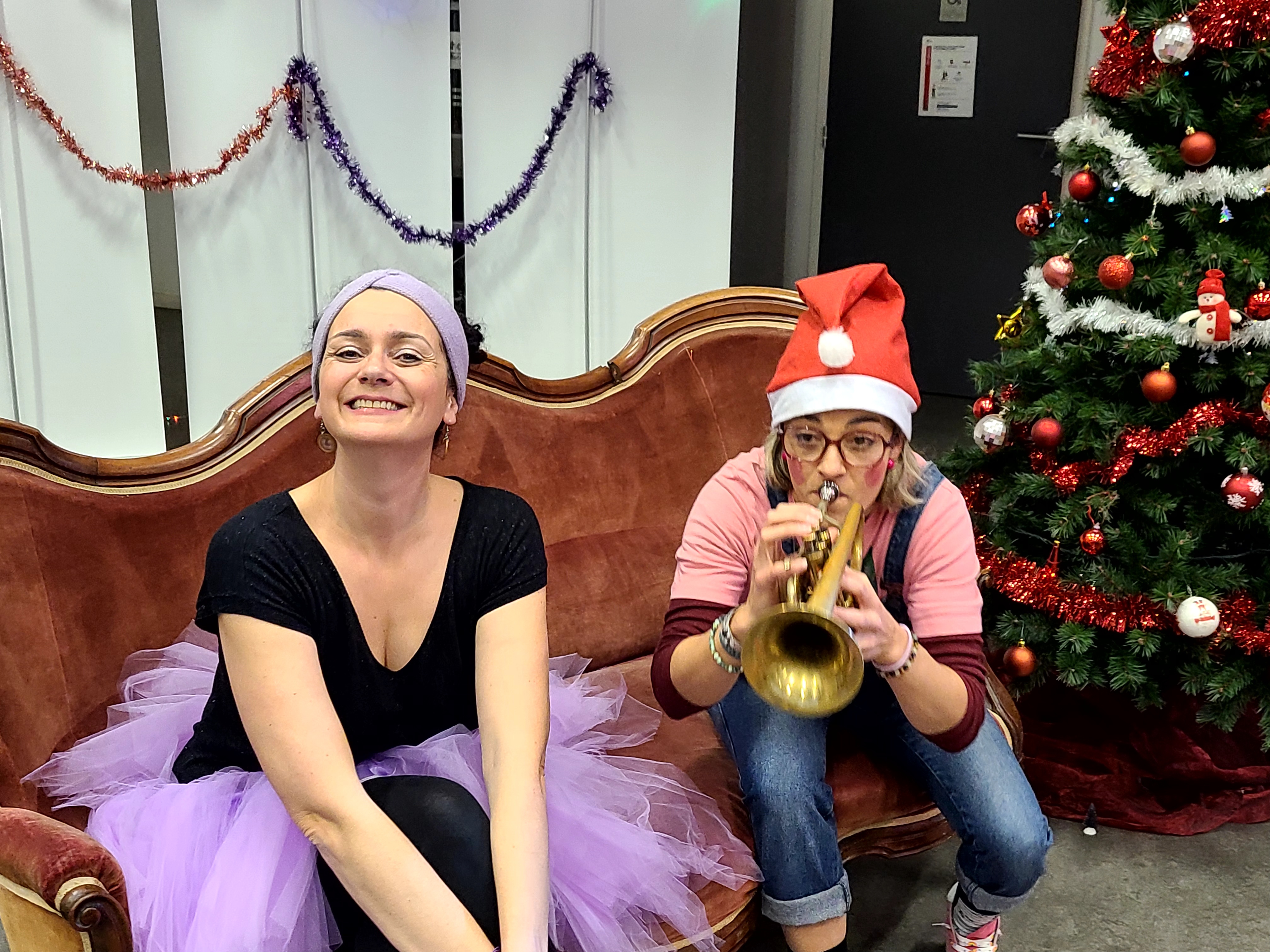 Sur le canapé, l'une des comédiennes sourit en tutu lilas pendant que l'autre joue de la trompette sous son bonnet de Père Noël.
