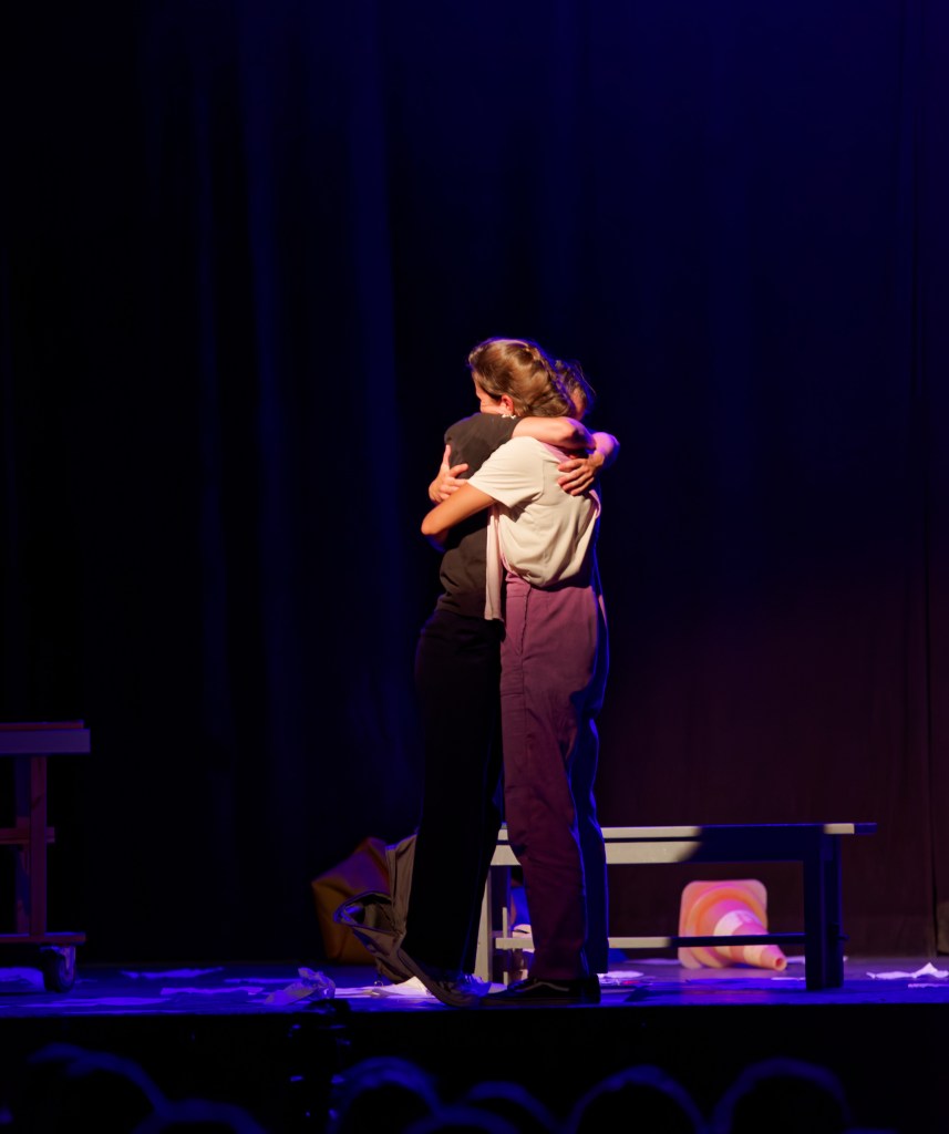 Les deux comédiennes s'enlacent sur scène, sous une lumière violette.